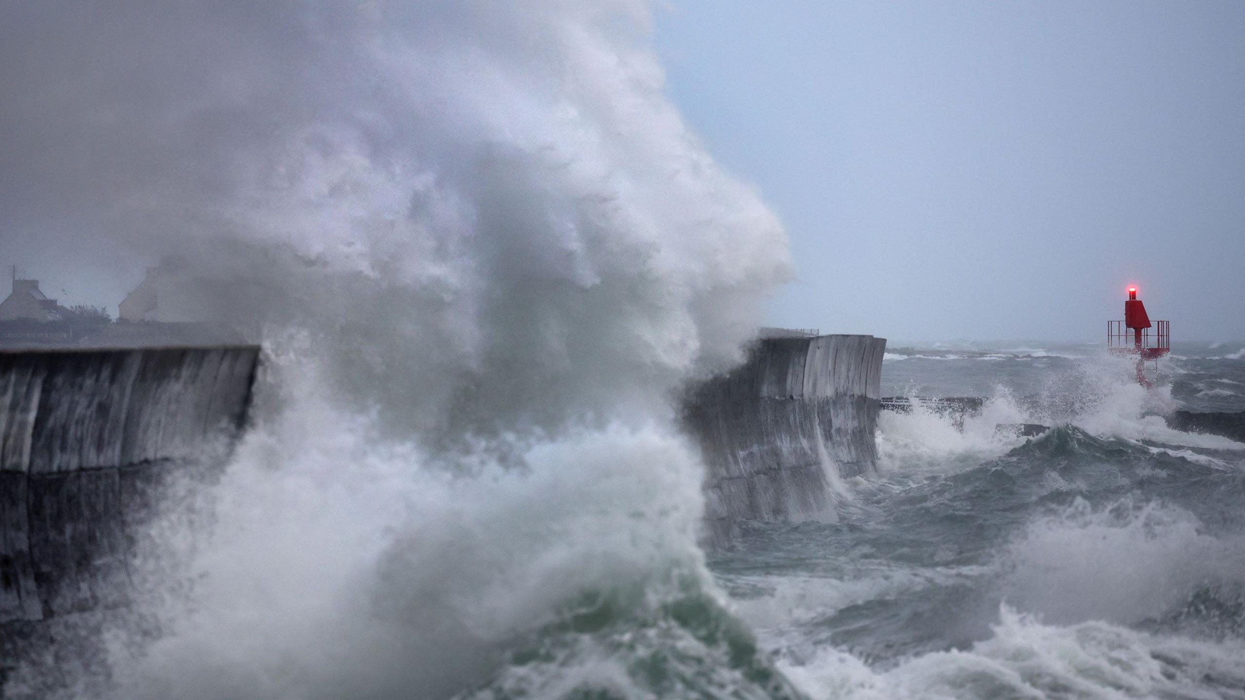 Massive waves crash against a pier on the UK coastline during stormy weather.