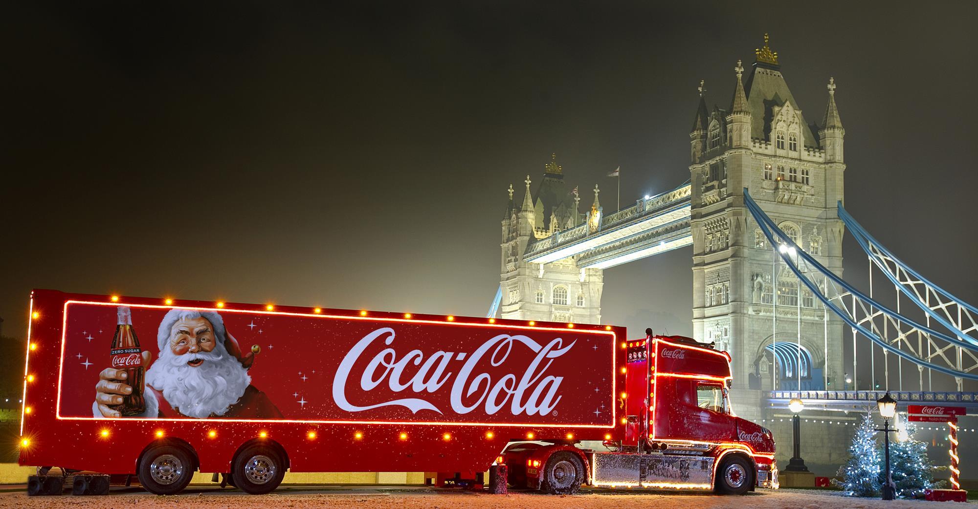 Iconic Coca-Cola holiday truck parked near Tower Bridge in London, lit up with festive red and white branding against the evening skyline.