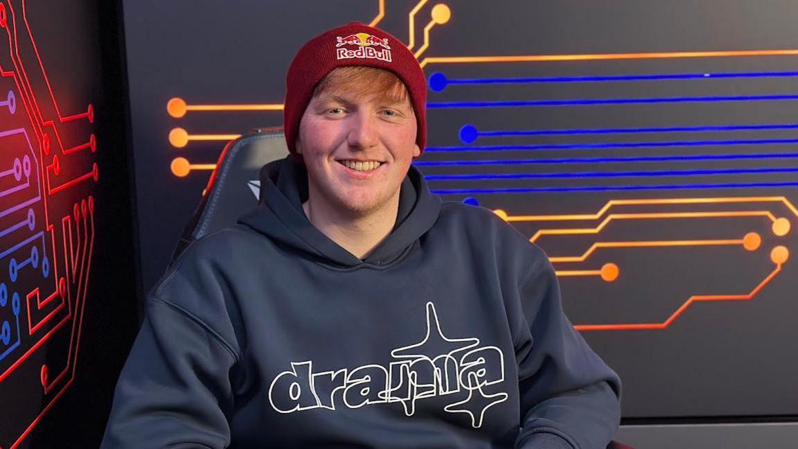 AngryGinge smiling behind a desk at a Red Bull event, wearing casual clothes and engaging with fans.
