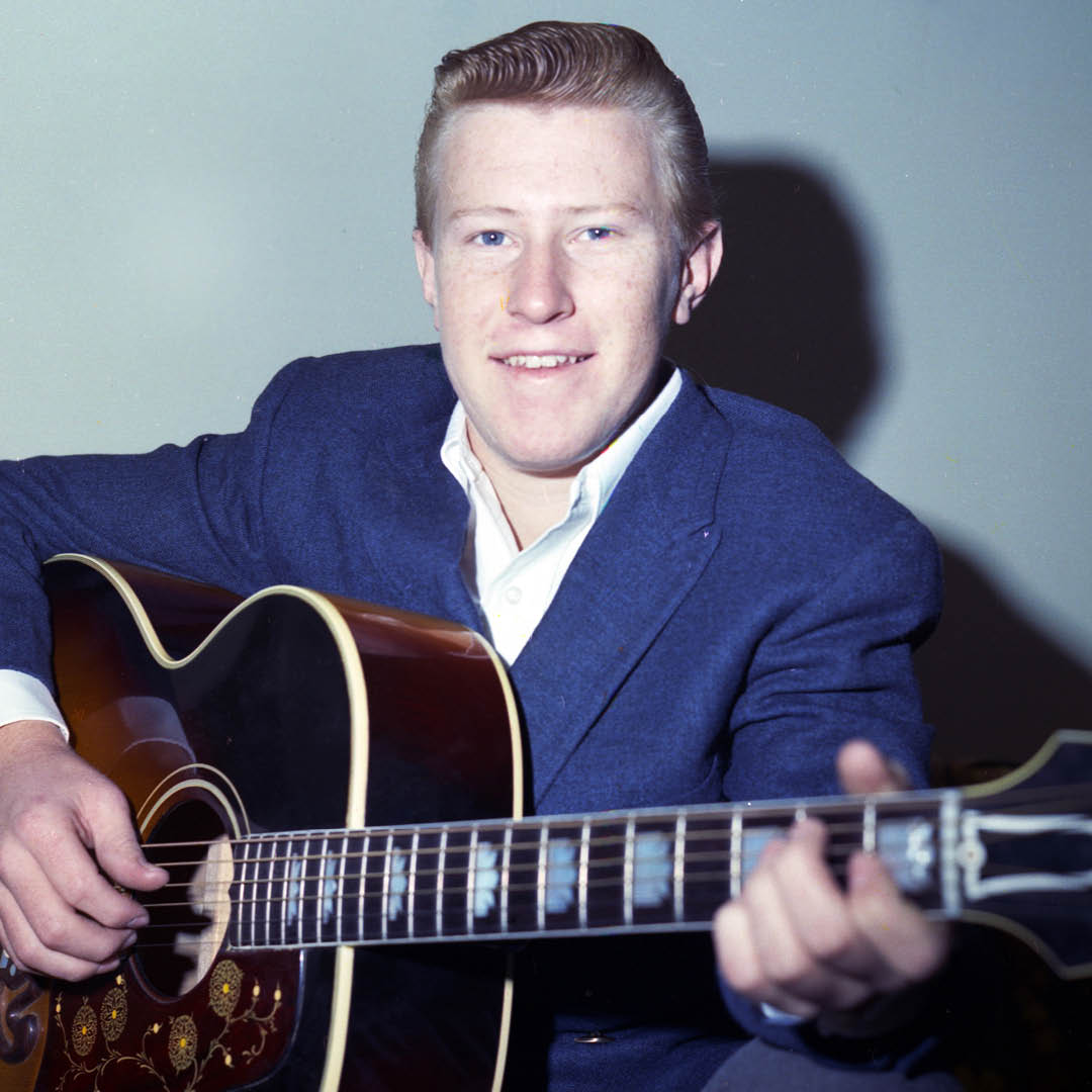 John Wesley Ryles posing in a blue suit while holding a guitar, smiling for the camera.