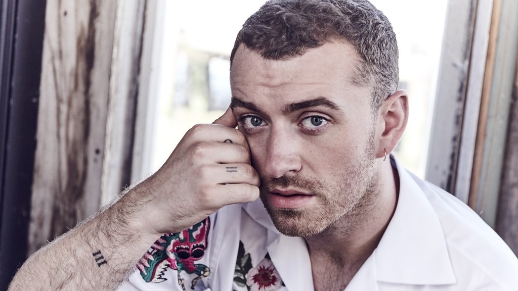 Sam Smith poses during a photoshoot in front of a large window, natural light illuminating their face as they look thoughtfully toward the camera.