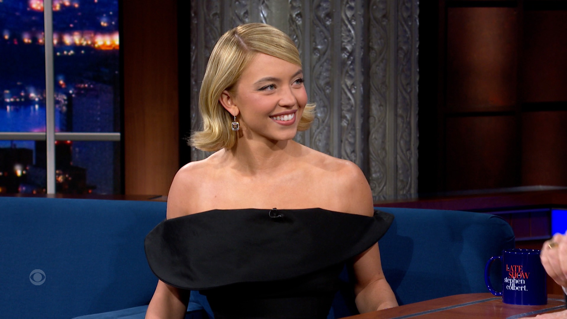 Sydney Sweeney sitting on The Late Show with Stephen Colbert sofa, animatedly telling the story of her skydiving adventure while smiling and gesturing.