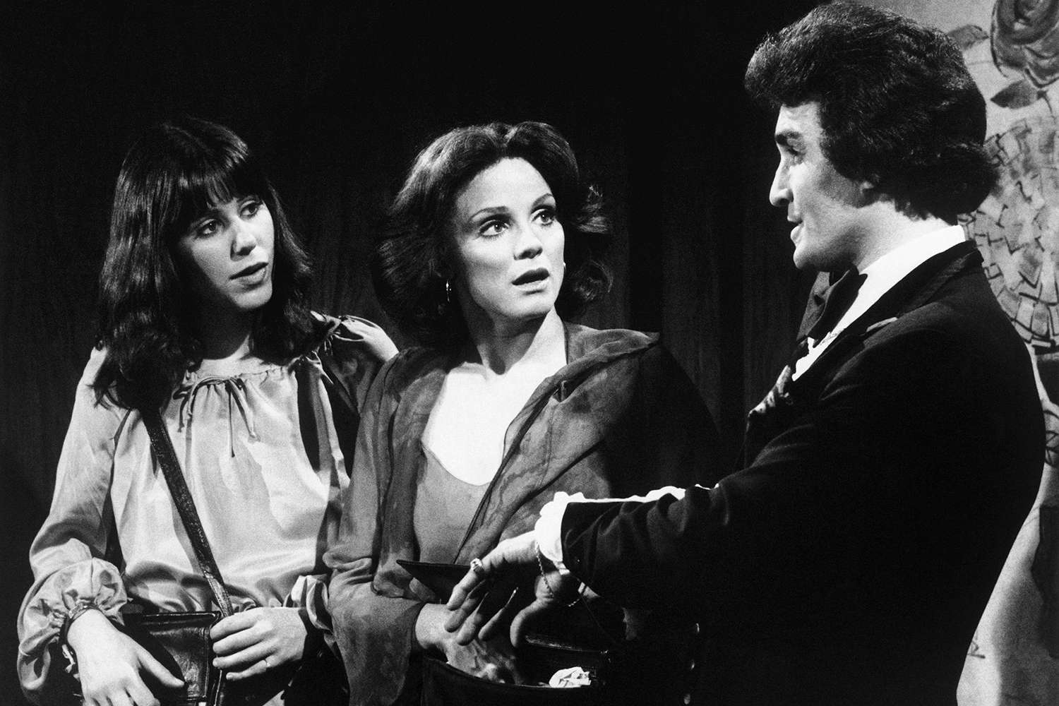 Black-and-white photo of Michael DeLano performing on stage, standing between two female singers, all dressed in classic performance attire.