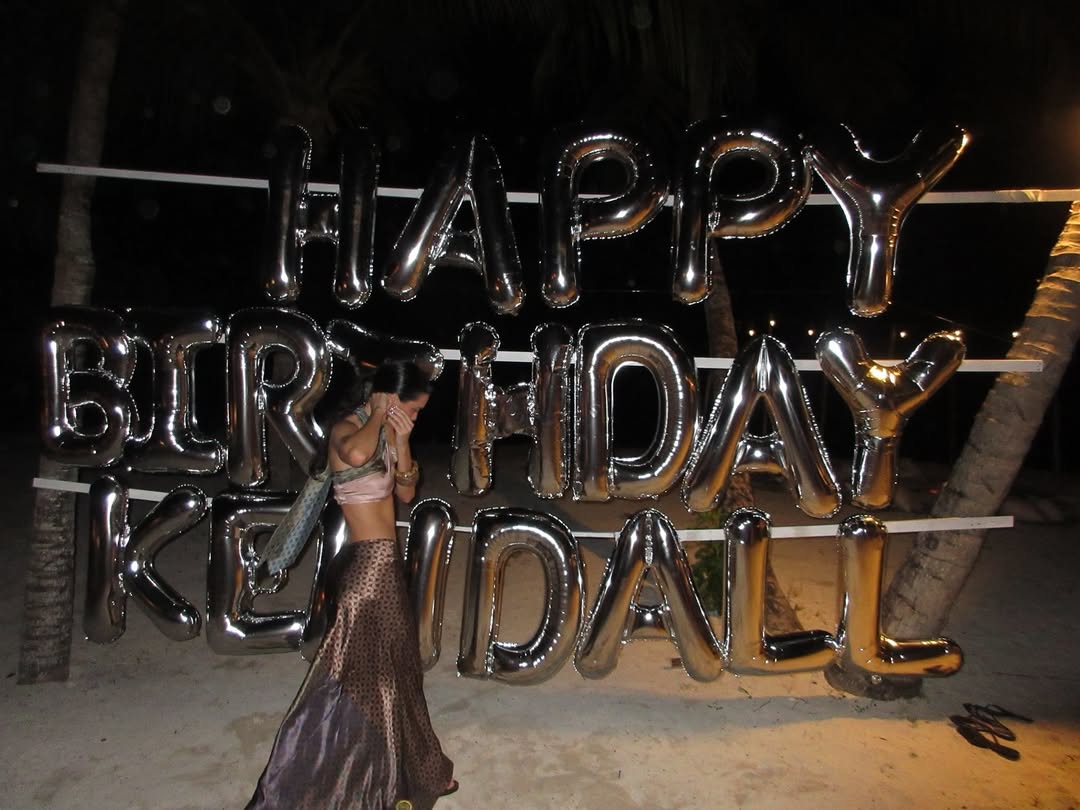 Kendall Jenner stands on the beach in front of silver “Happy Birthday Kendall” balloons, wearing a pink halterneck crop top and patterned wrap skirt, smiling at the camera.