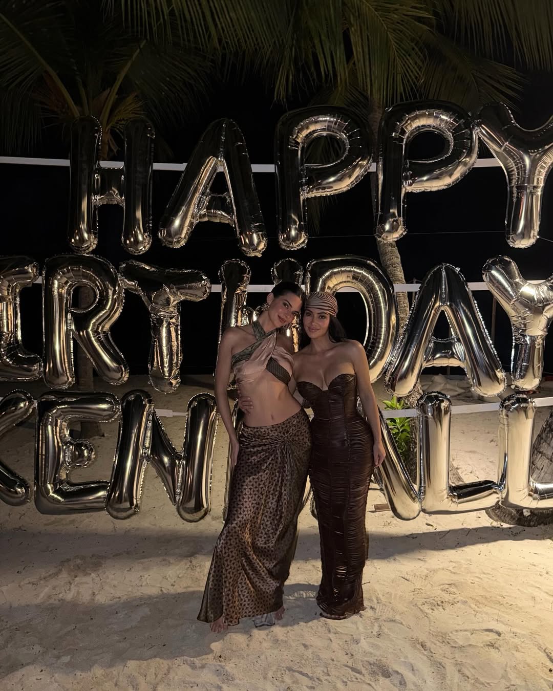 Kim Kardashian and Kendall Jenner posing together in front of a large silver balloon display spelling “Happy Birthday Kendall,” both dressed in revealing outfits.