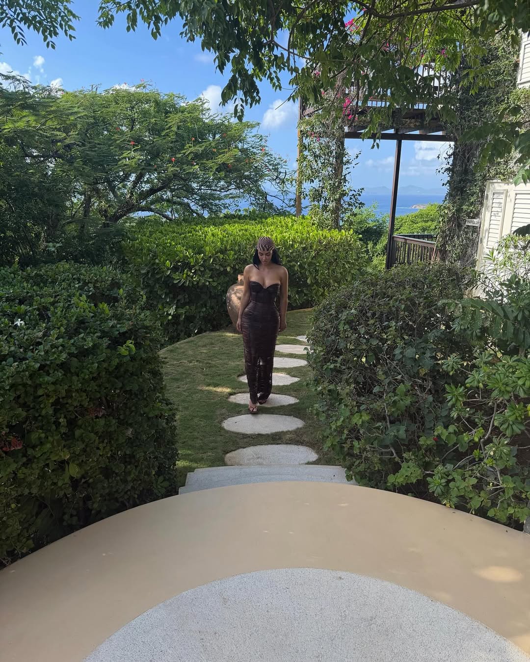 Kim Kardashian walking gracefully through lush tropical gardens wearing a brown off-the-shoulder shredded dress, oversized sunglasses, and black open-toe heels.