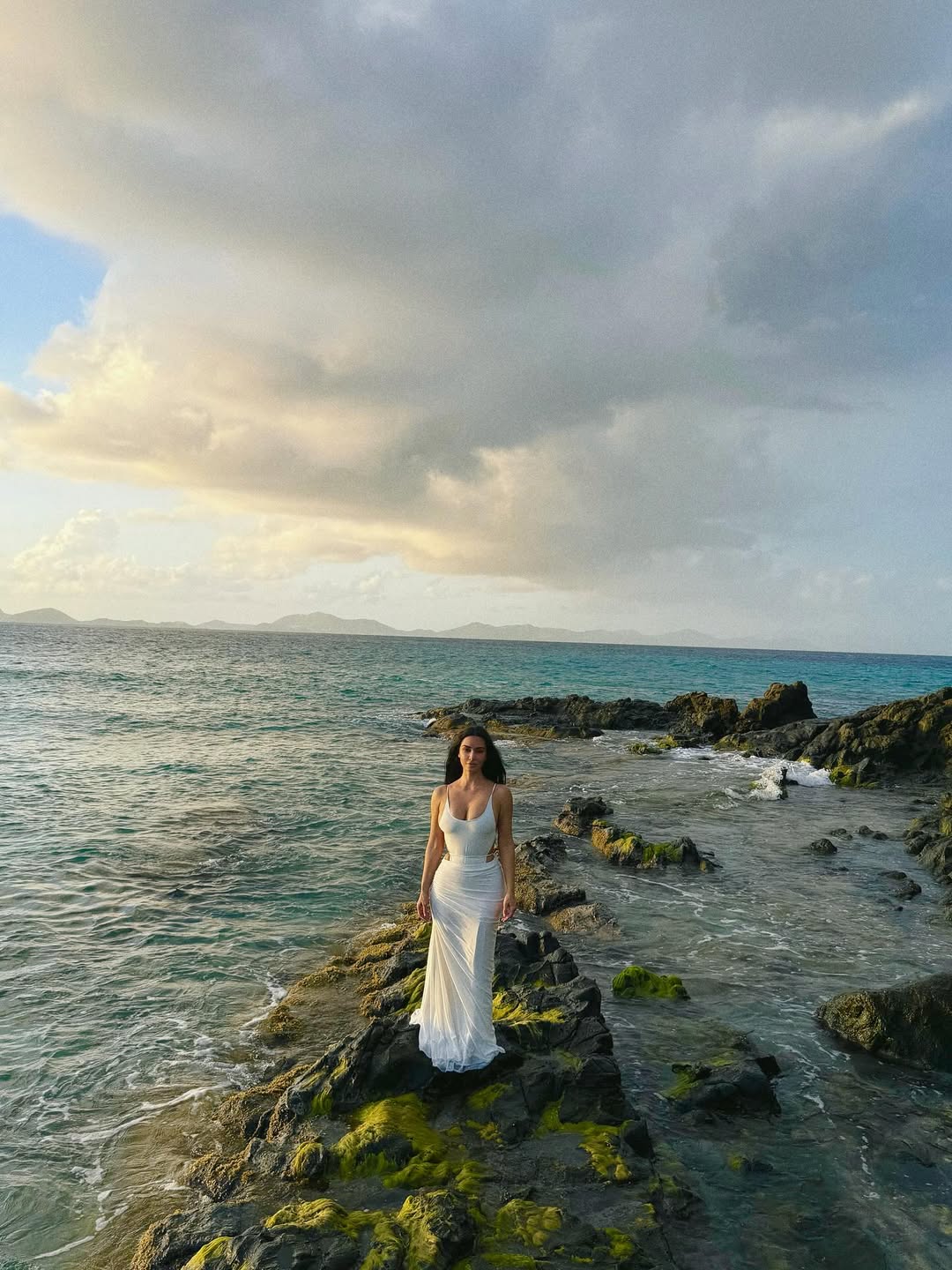 Kim Kardashian perched on a rock surrounded by the sea, wearing a flowing white dress, exuding elegance and calm on bar exam results day.
