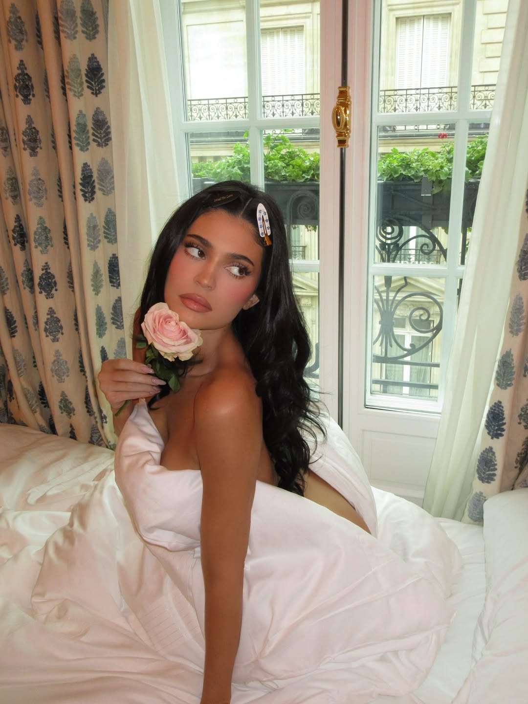 Kylie Jenner sitting nude in bed with a duvet wrapped around her body, holding a pink rose while looking away from the camera.