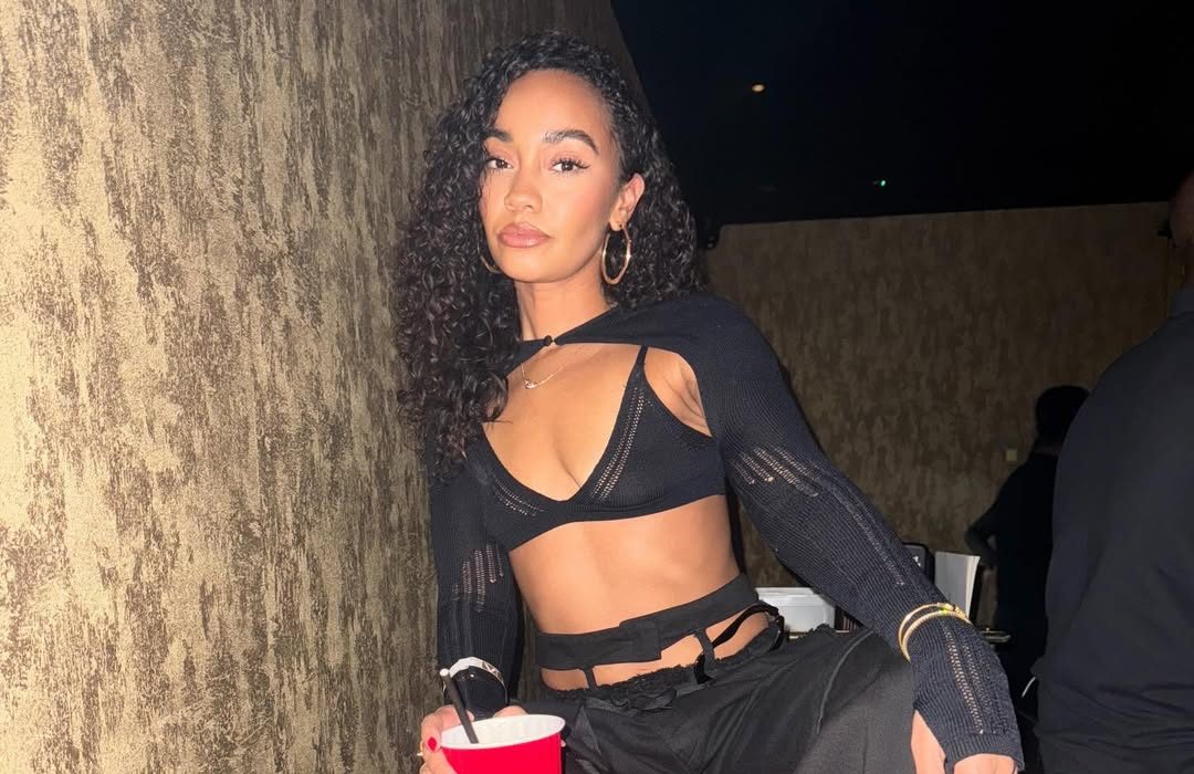 Leigh-Anne Pinnock poses confidently in a black bra-top outfit, holding a red party cup and giving a bold, rebellious look to the camera.