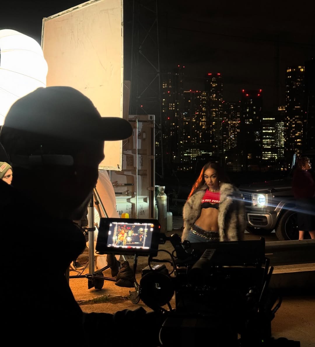 Leigh-Anne Pinnock poses for a sultry hotel-room photoshoot, standing by the window in her sleek bra-top outfit with a glowing city skyline behind her.