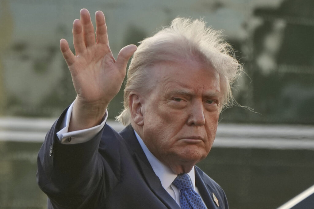 Donald Trump looking serious and glum while raising his hand in a wave, dressed in a dark suit.