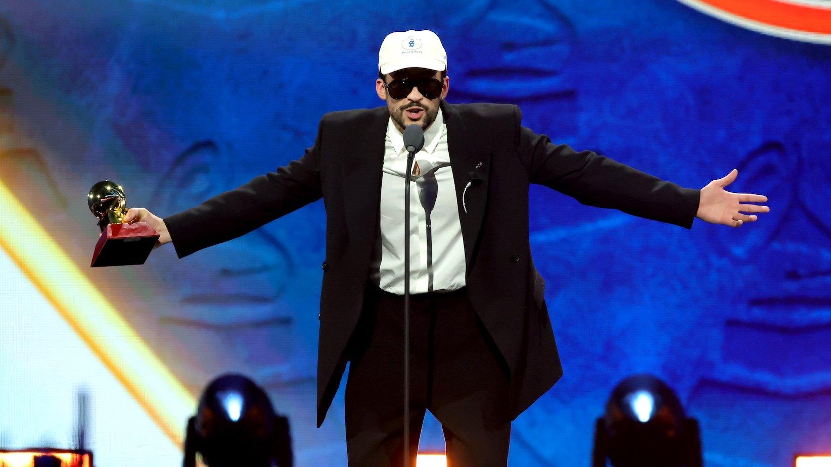 Bad Bunny on stage holding a Latin Grammy trophy, speaking into a microphone, wearing a colorful outfit, with a spotlight behind him.