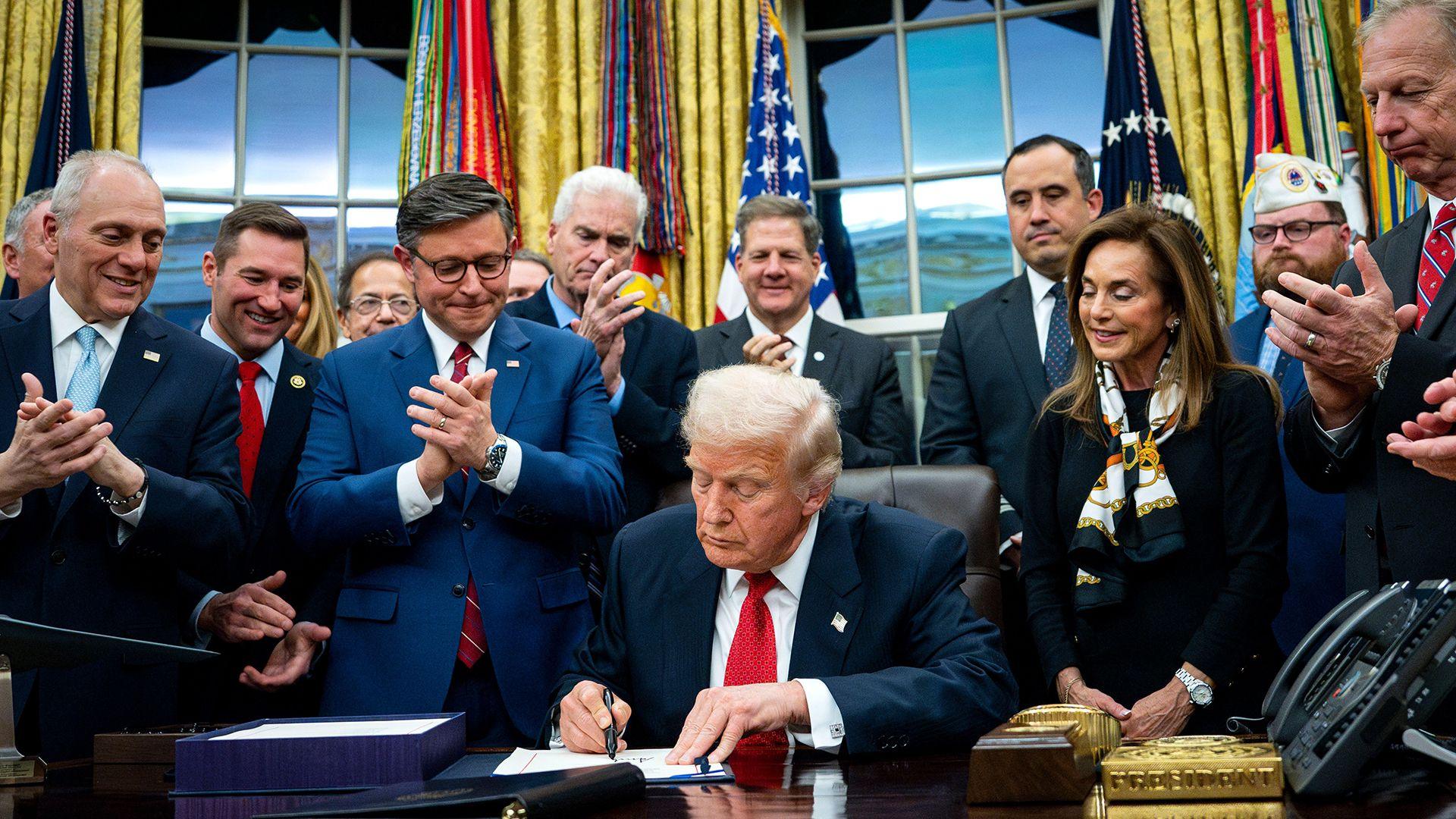 U.S. President Donald Trump sitting at a desk, signing the 2025 federal spending bill into law with an American flag in the background.