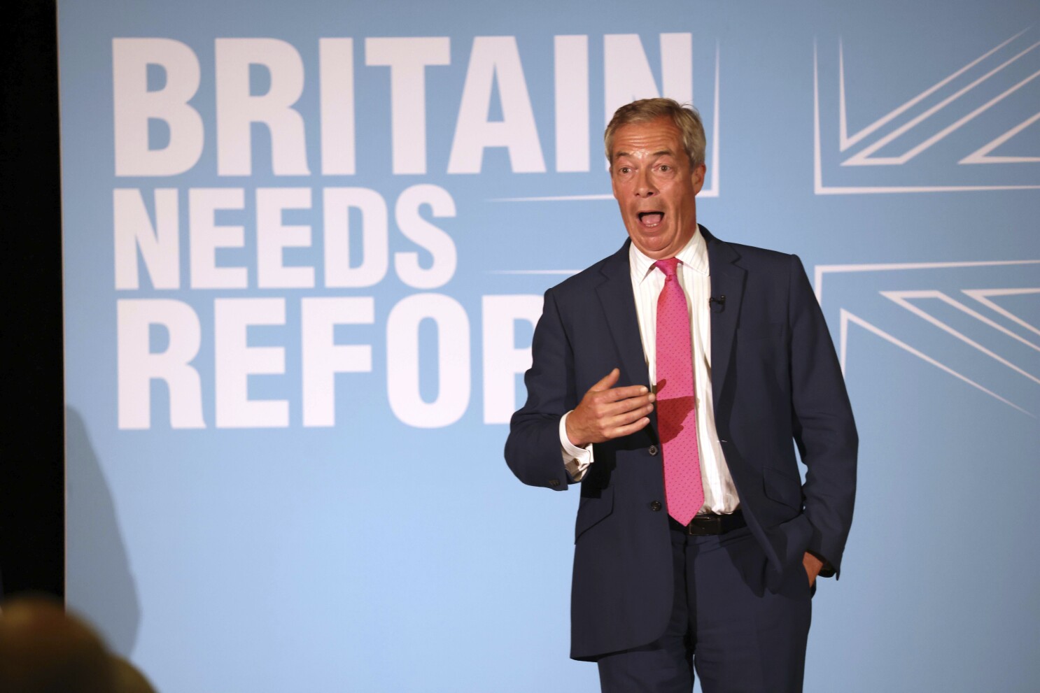 Nigel Farage speaking passionately on stage at a rally, gesturing emphatically while delivering his signature combative and polarizing rhetoric.