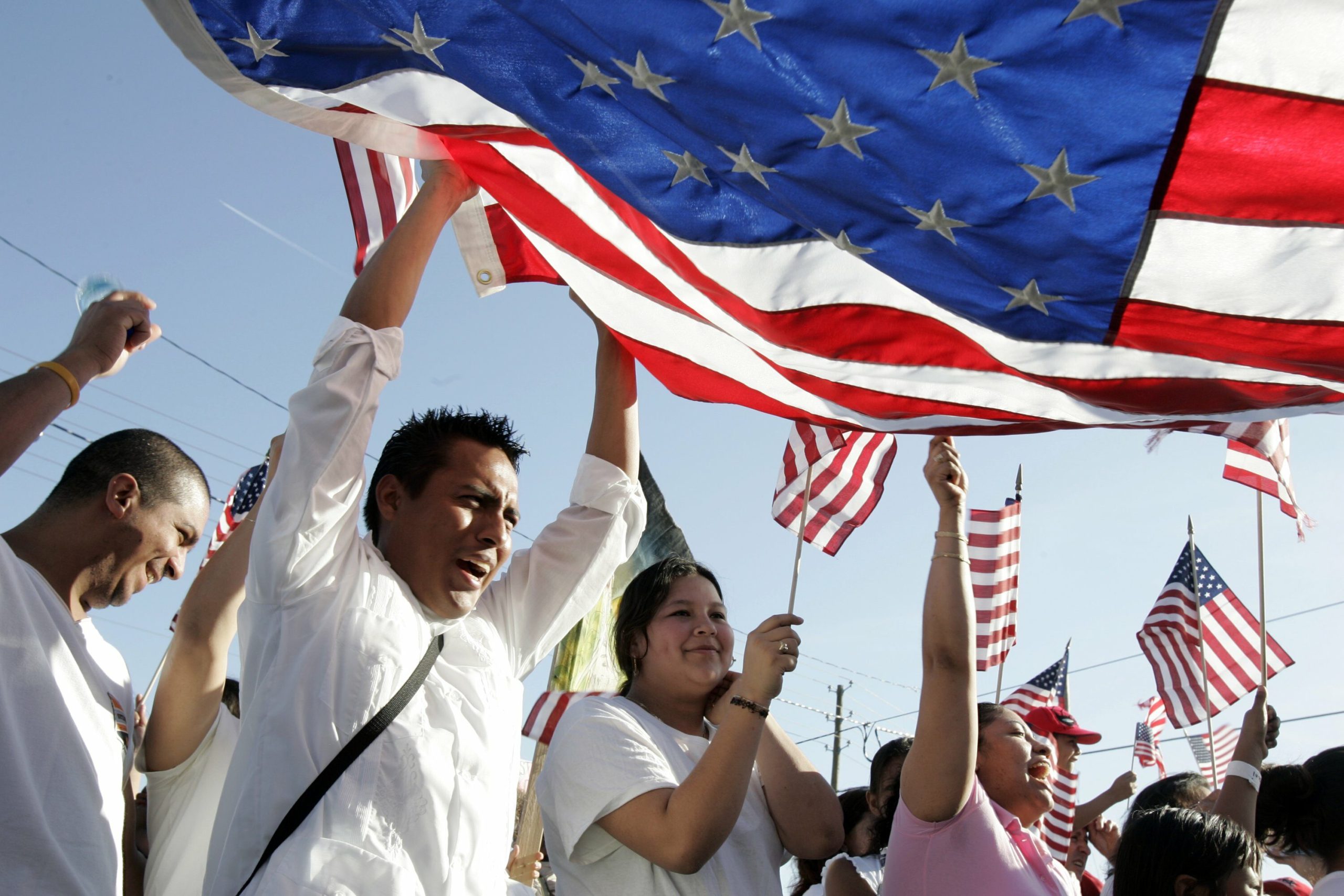 A group of immigrants smiling and waving small American flags during a citizenship or immigration event, reflecting hope and inclusion amid changing national policies.
