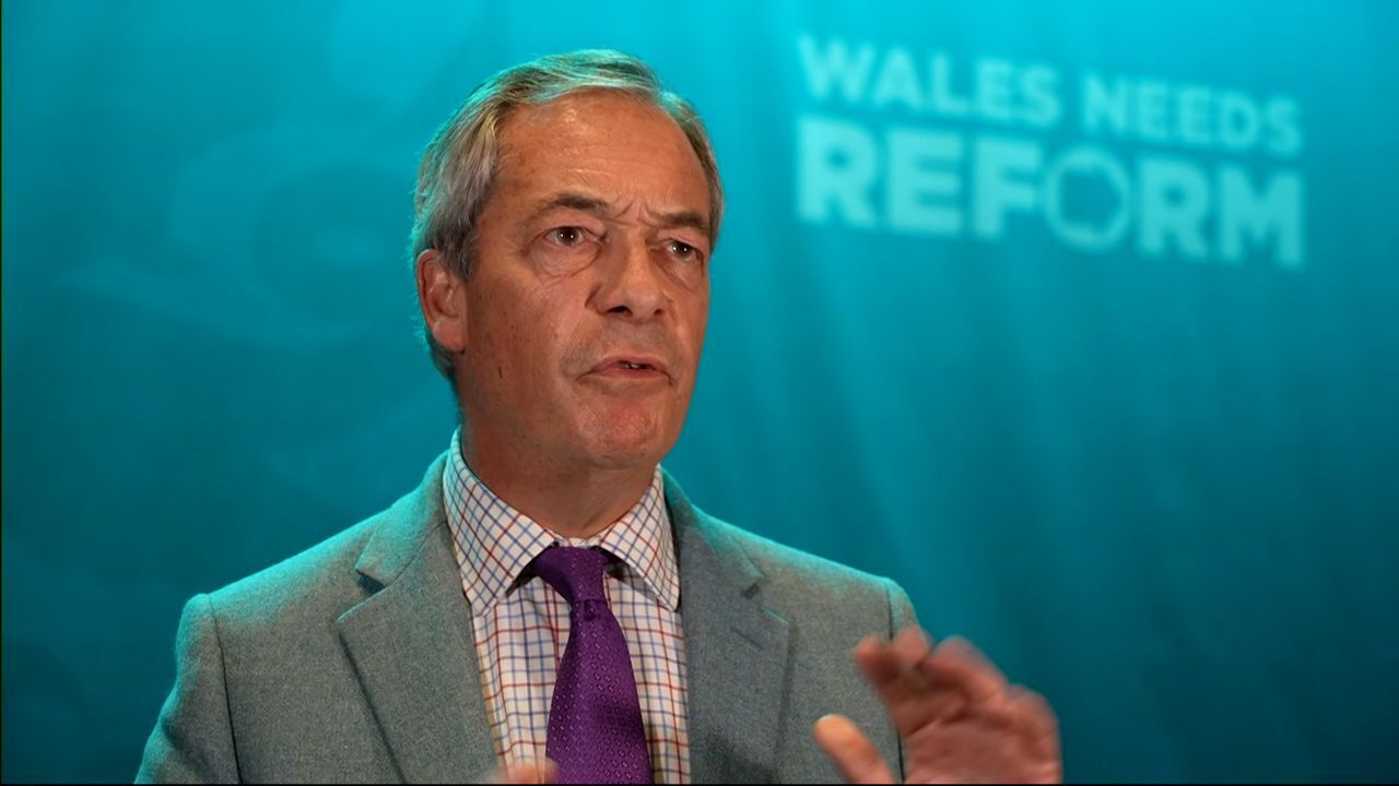 Nigel Farage speaking during a televised interview, gesturing with his hands while addressing allegations, looking composed yet serious.