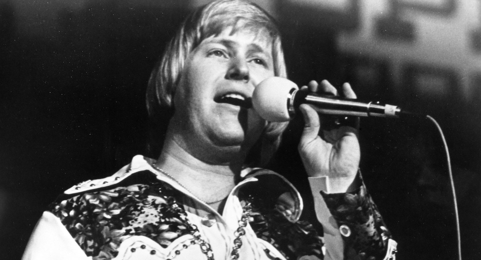 Black and white photo of country music legend John Wesley Ryles passionately performing on stage, holding a microphone.