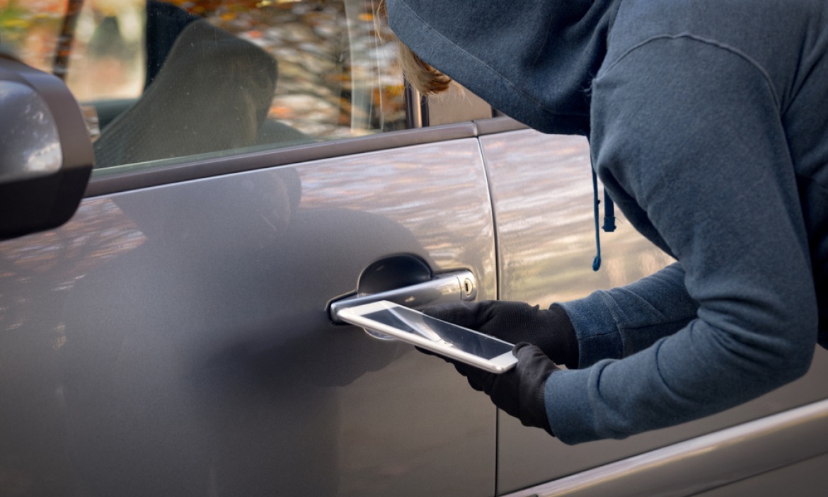 A masked car thief using an electronic relay device beside a car door handle during a keyless vehicle theft.