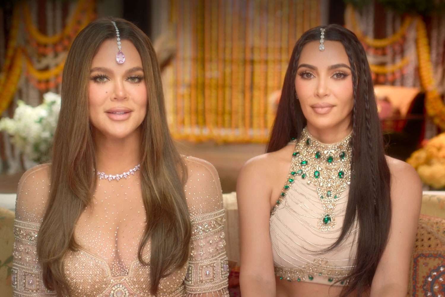Khloe Kardashian and Kim Kardashian sitting together, fully glammed up in sleek evening looks, sharing a knowing smile that hints at their sisterly bond and the family history resurfacing with the return of their late father’s Bible.