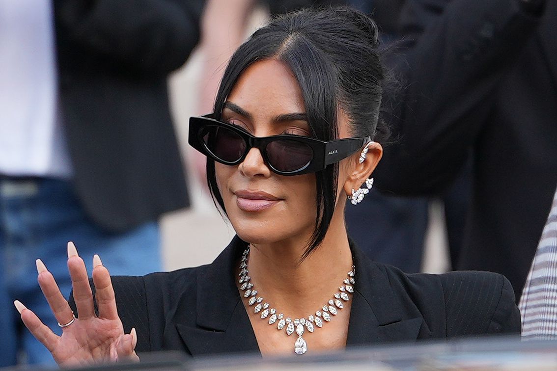 Kim Kardashian waving to photographers outside the Paris courthouse, wearing a sleek black blazer and showcasing her multi-million-dollar diamond necklace and earrings.