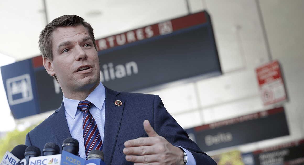 Eric Swalwell speaking to a group of reporters, microphones clustered around him as he delivers focused remarks during his campaign for California governor.