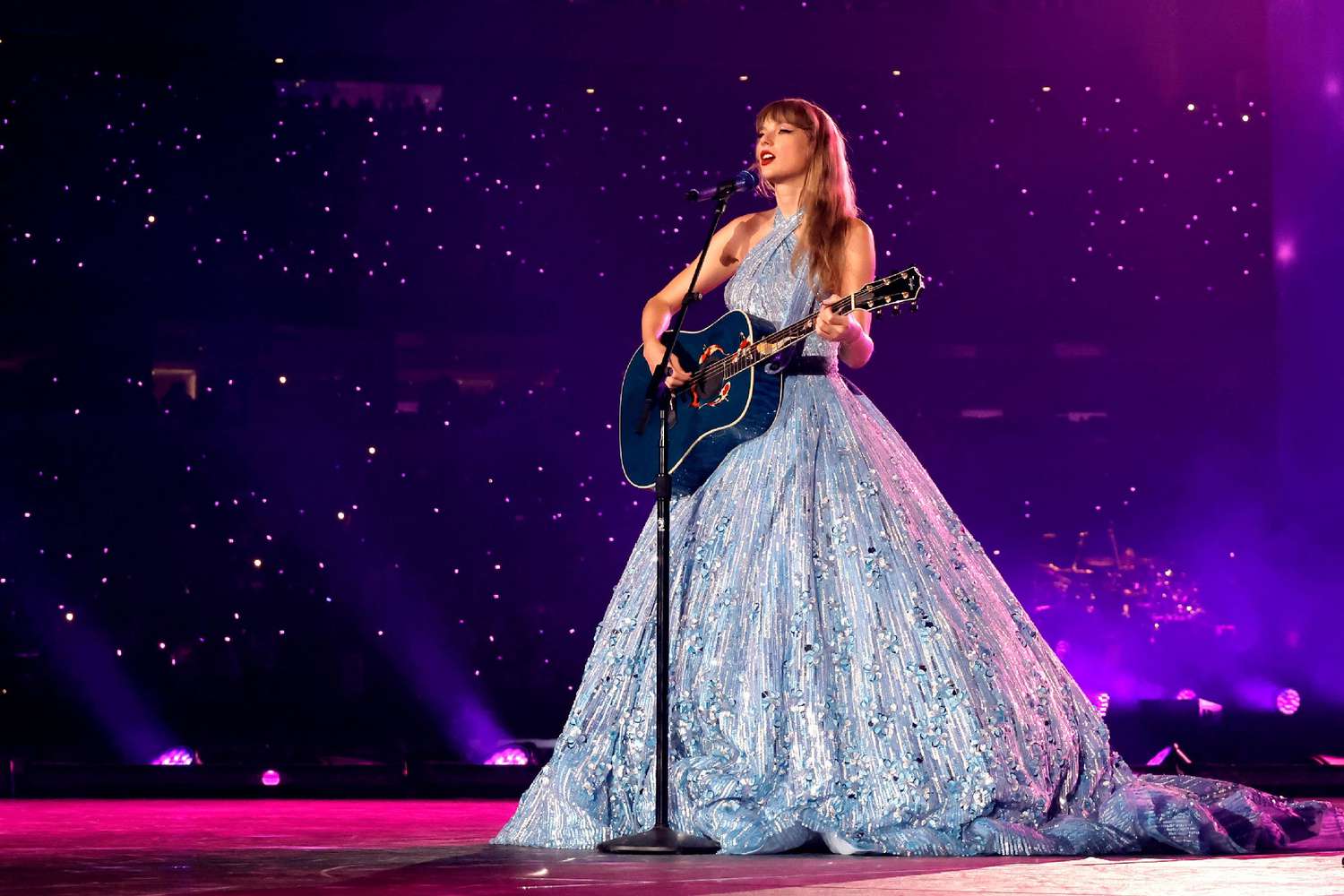 Taylor Swift performing on the Eras Tour in a long, flowing blue dress while playing an acoustic guitar, highlighting a signature moment from the tour captured in her upcoming Disney+ docuseries.