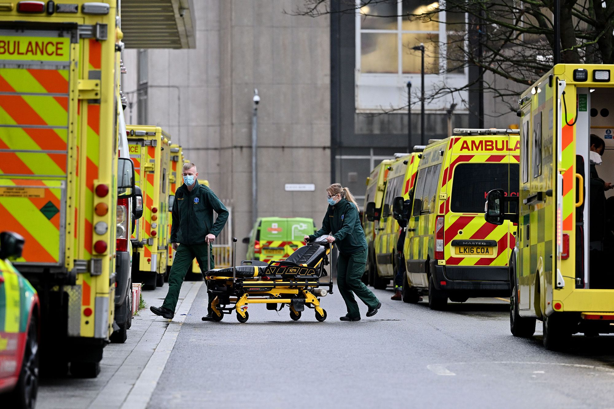 A line of ambulances parked outside a busy hospital emergency entrance amid high patient demand.