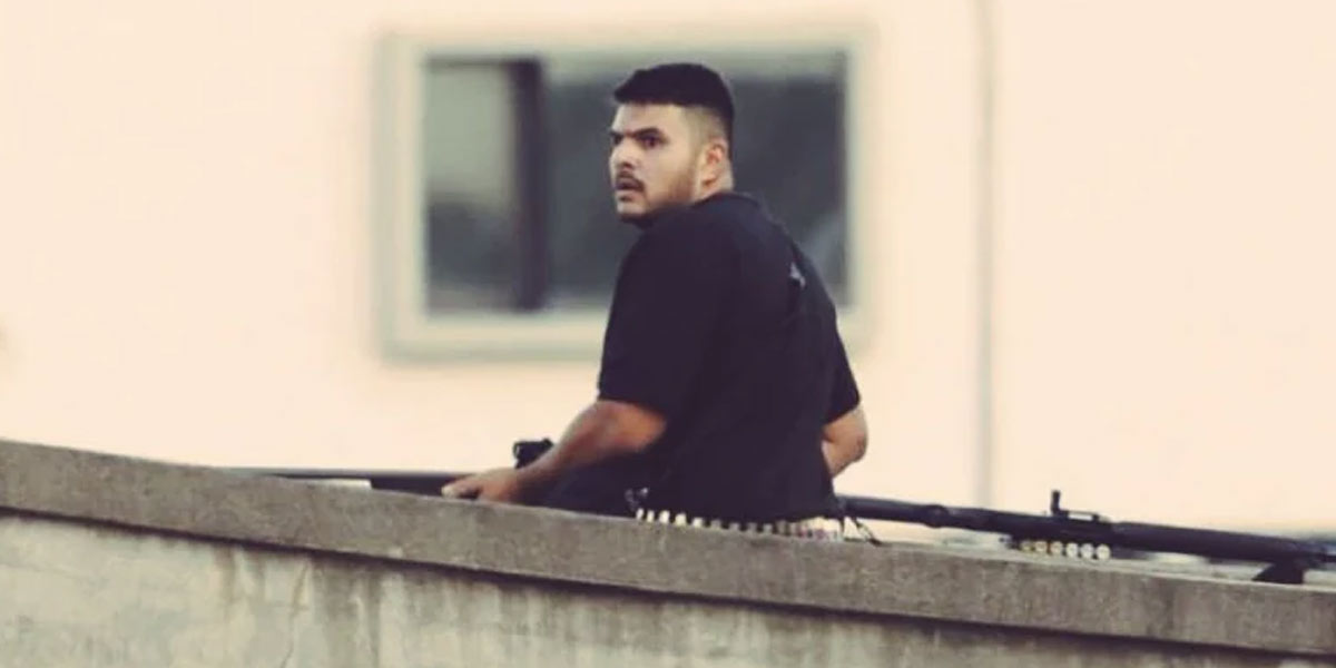 Alt Text A blurred or distant image from verified footage showing suspected shooter Naveed Akram, dressed in dark clothing, positioned on a pedestrian footbridge overlooking Bondi Beach during the December 2025 terrorist attack, appearing to aim and fire a rifle toward crowds below.