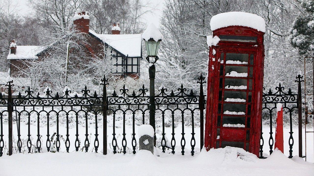 A traditional red British phone box standing in fresh snow during a white Christmas scene, surrounded by softly falling snowflakes and glowing streetlights in a quiet winter setting.