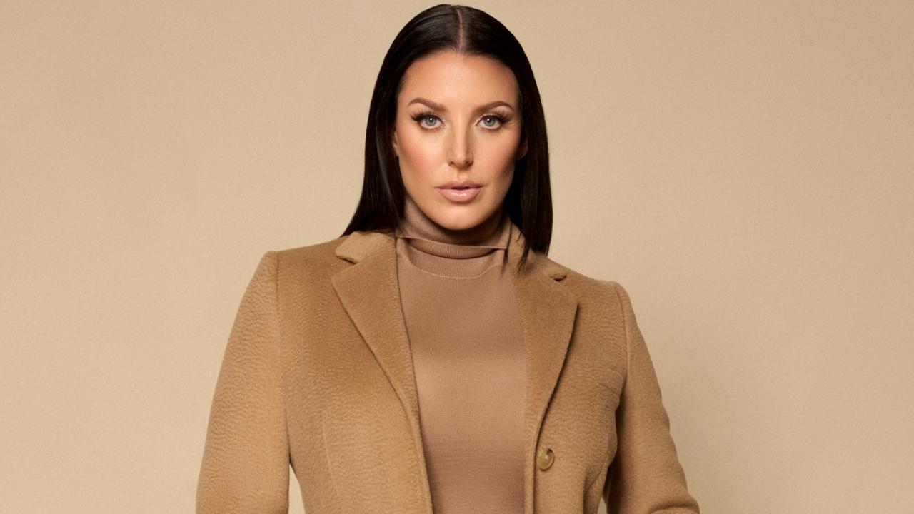 Angela White posing in a beige outfit during a professional photoshoot, looking confident as she stands against a soft studio backdrop.