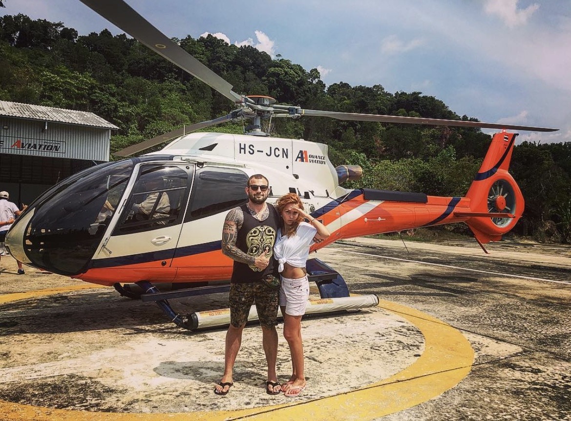 Roman Novak and his wife Anna standing in front of a helicopter, dressed casually and posing for a photo, exuding a luxurious lifestyle.