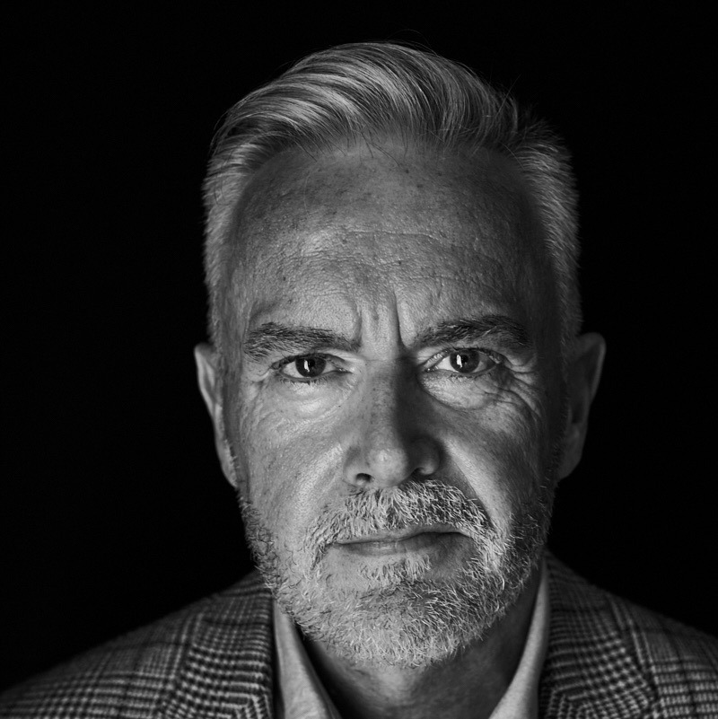 Professional black-and-white portrait of Huw Edwards in a suit jacket, with white stubble and a contemplative expression.