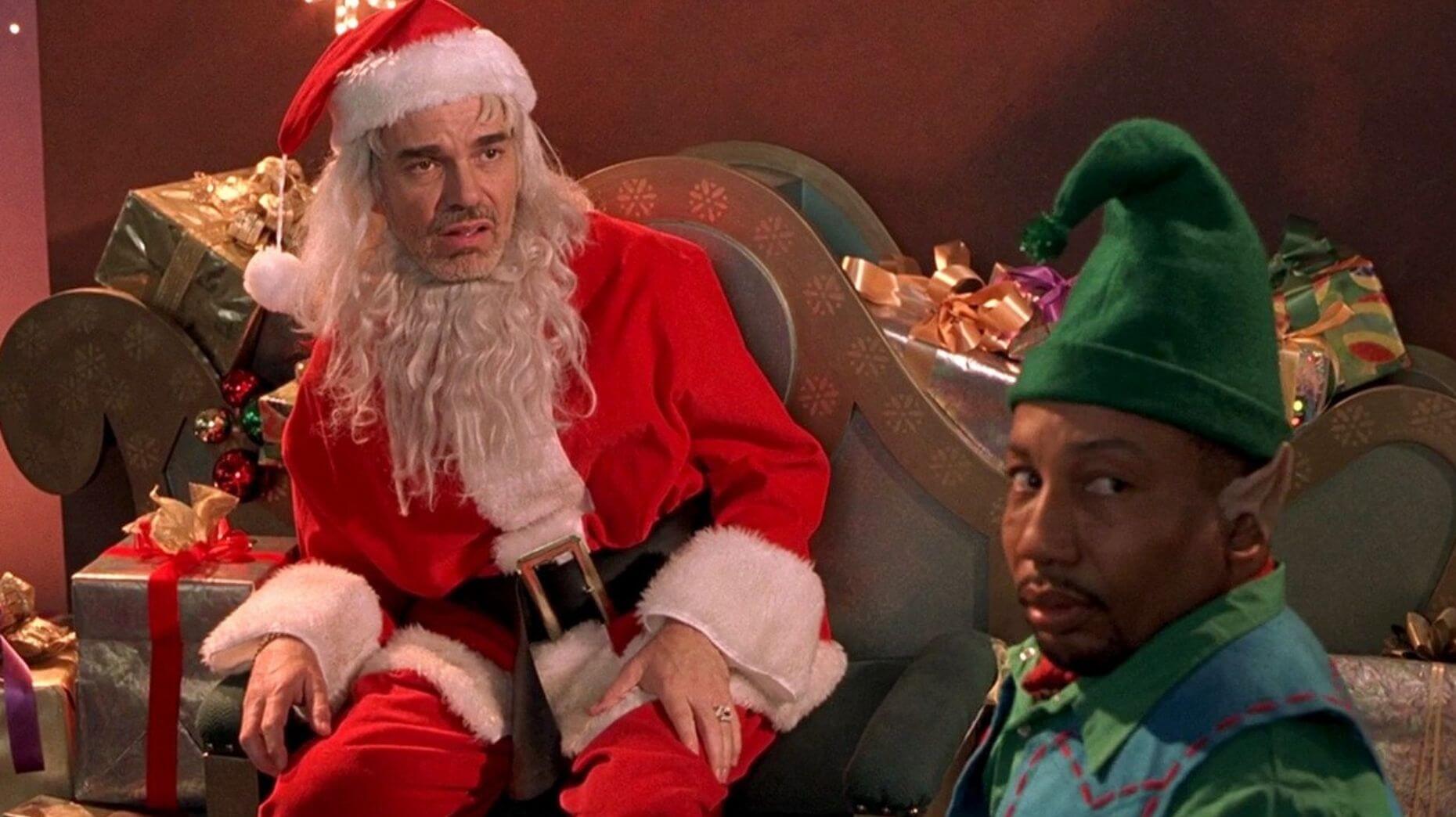 Billy Bob Thornton in full Bad Santa costume, looking grumpy and disheveled with a cigarette dangling from his mouth, embodying the film’s iconic anti-hero.