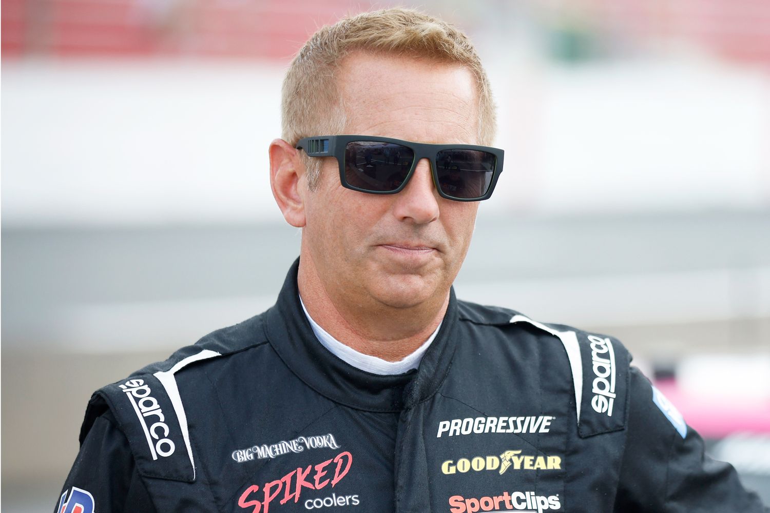 Greg Biffle wearing his NASCAR racing overalls and sunglasses, standing confidently in the pit area before a race.