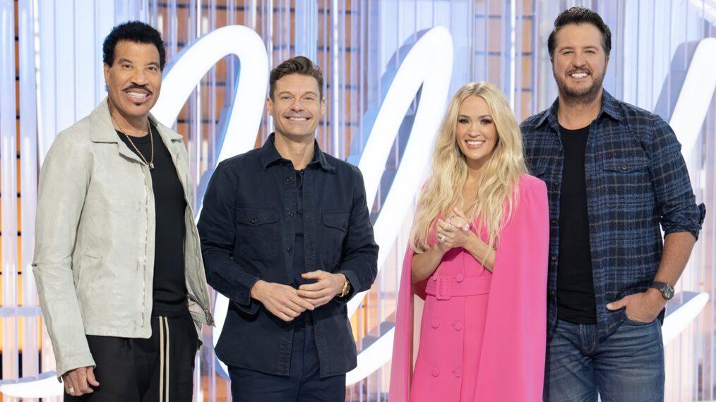 The 2025 American Idol judging panel seated on stage, watching contestants perform, with microphones in front of them and studio lights highlighting their expressions.