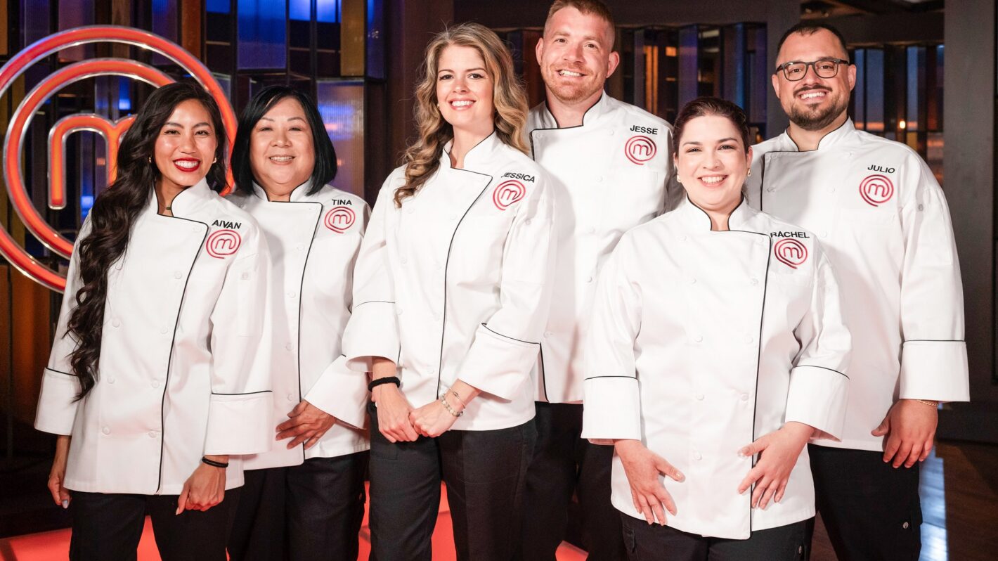 The 2025 cast of MasterChef USA standing together in the competition kitchen, wearing aprons and smiling confidently at the camera.