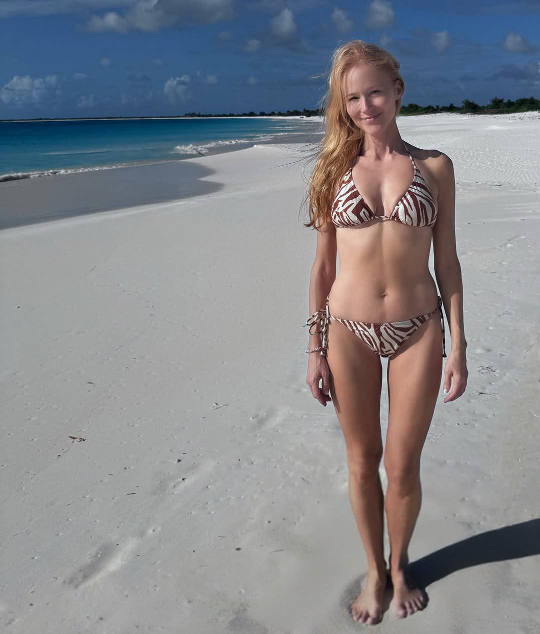 Jewel posing on a sun-soaked beach in a tiny brown-and-tan zebra-print bikini, showing her toned abs and muscular legs.