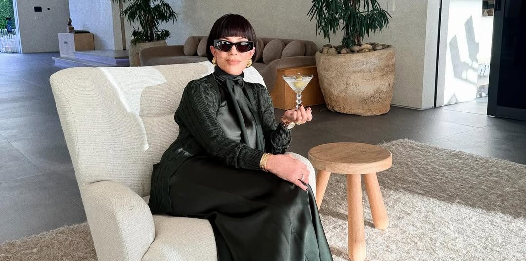 Kris Jenner posing with her sleek new bob hairstyle, holding a martini glass in one hand, wearing a forest-green outfit and dark sunglasses in a festive holiday setting.