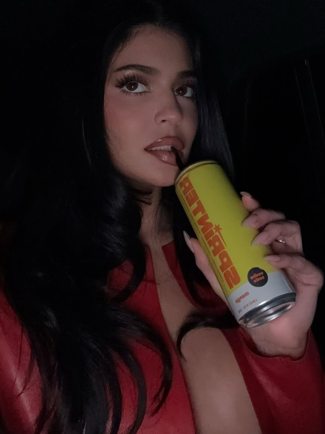 Kylie Jenner sipping a drink through a straw while wearing a plunging red leather cropped jacket and low-rise jeans, standing by a white Christmas tree with soft curls and festive glam.