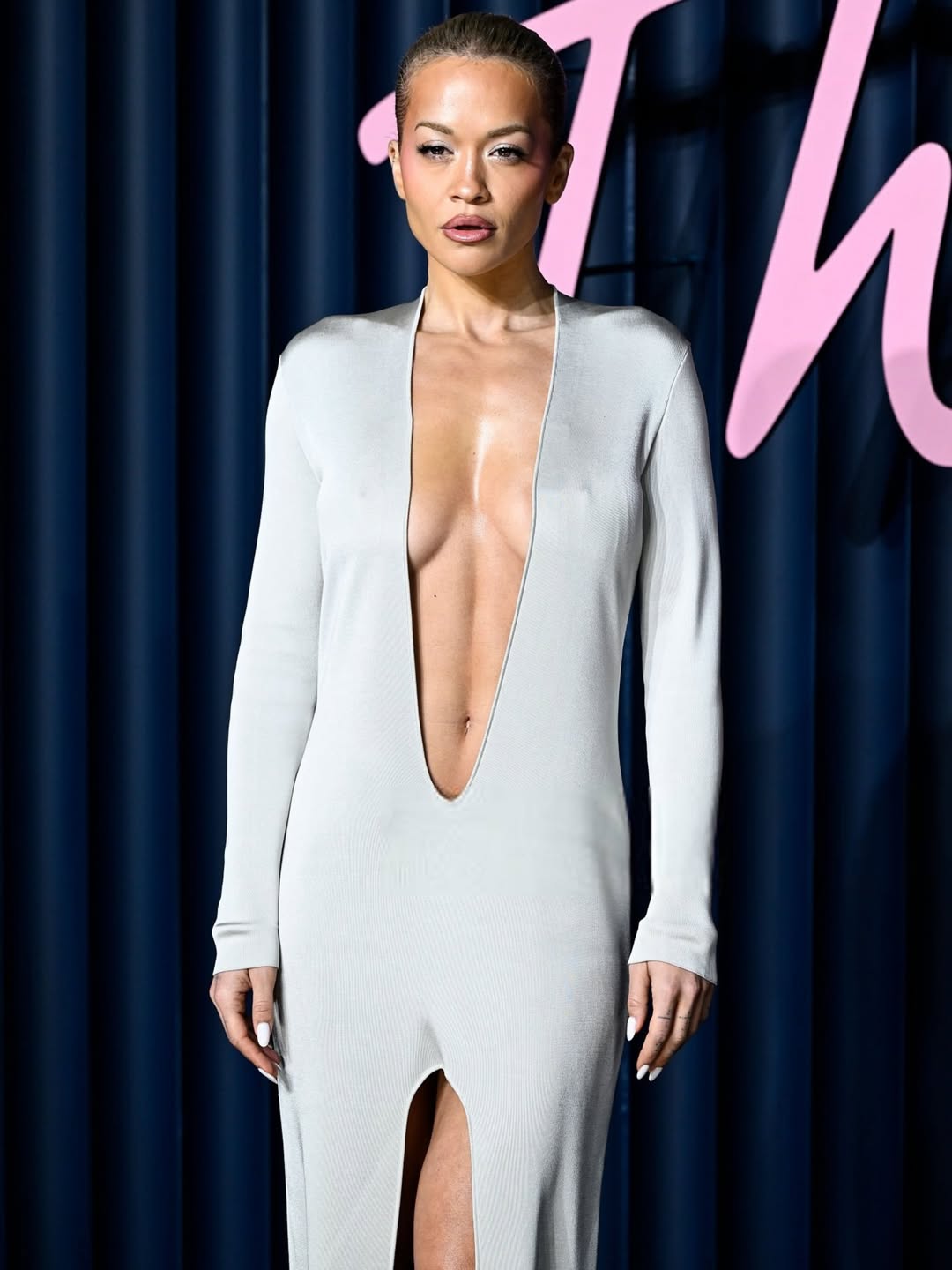 Rita Ora posing in full-length, showcasing her plunging silver gown on the Fashion Awards red carpet, highlighting the daring cut and sleek silhouette.