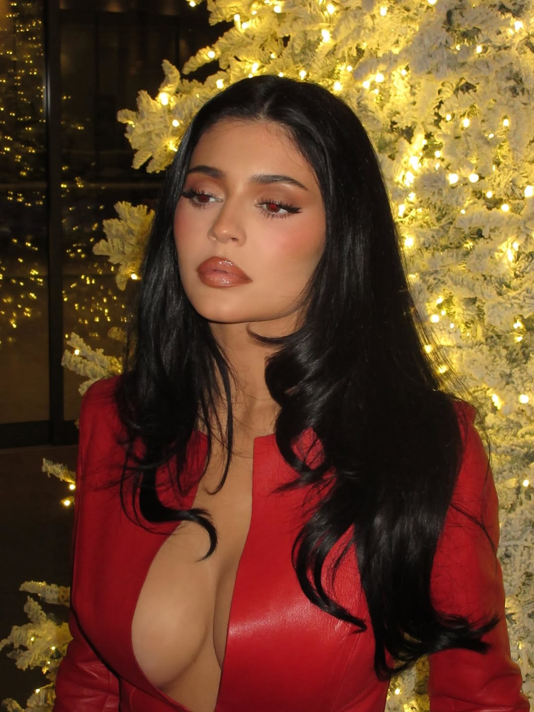 Kylie Jenner gazes off to the side while posing in a plunging red leather cropped jacket in front of a white Christmas tree, her long dark curls and cranberry-toned makeup completing the festive look.