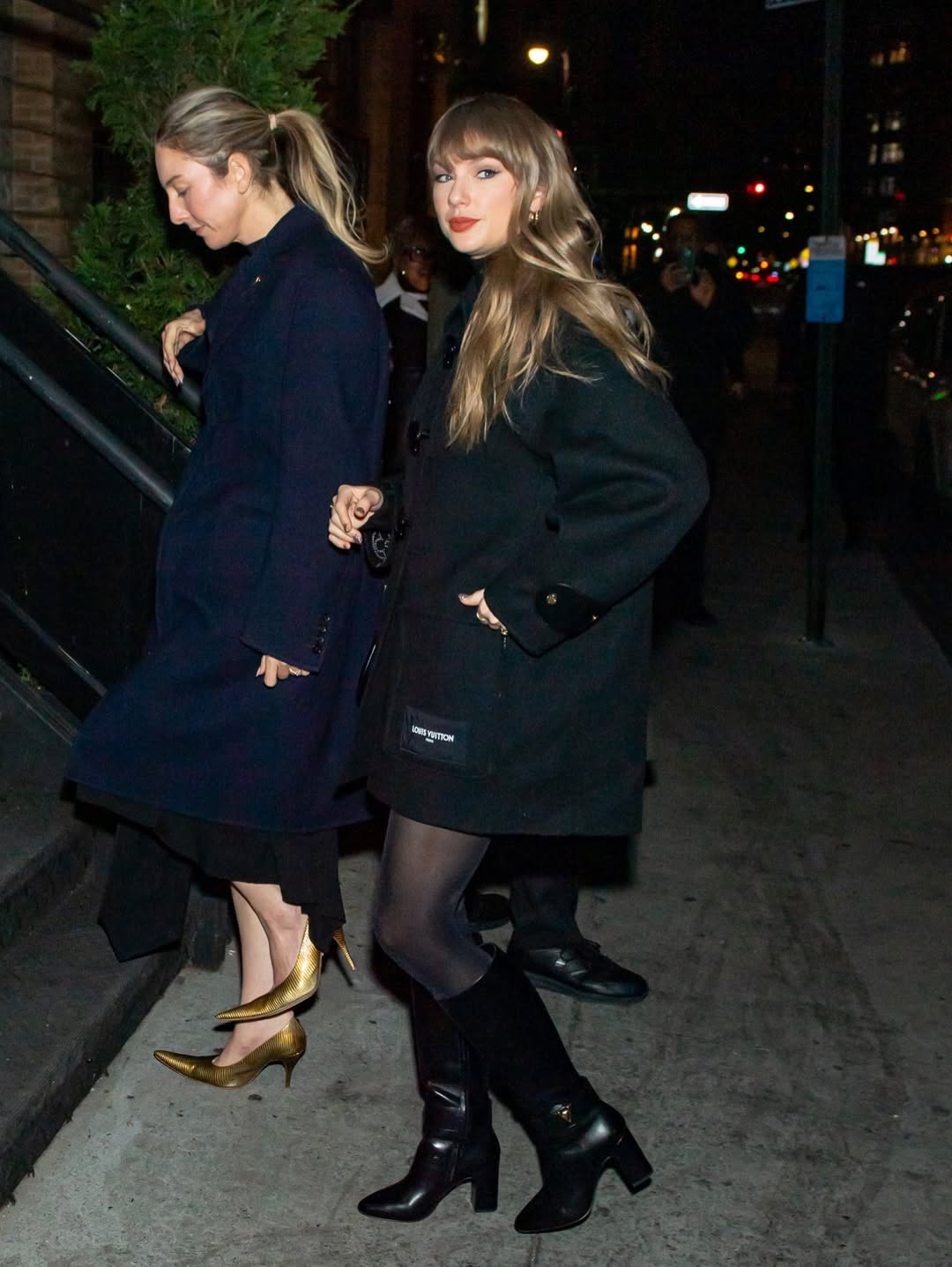 Full-length view of Taylor Swift in a black Louis Vuitton Double Face Duffle Coat, matching leather Midtown High Boots, and sheer black tights, standing on a NYC sidewalk at night.