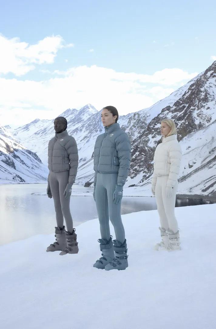 Three models wearing additional pieces from the SKIMS x The North Face 2.0 collection, including full-length ski suits, puffers, and accessories, posing on a mountain top.