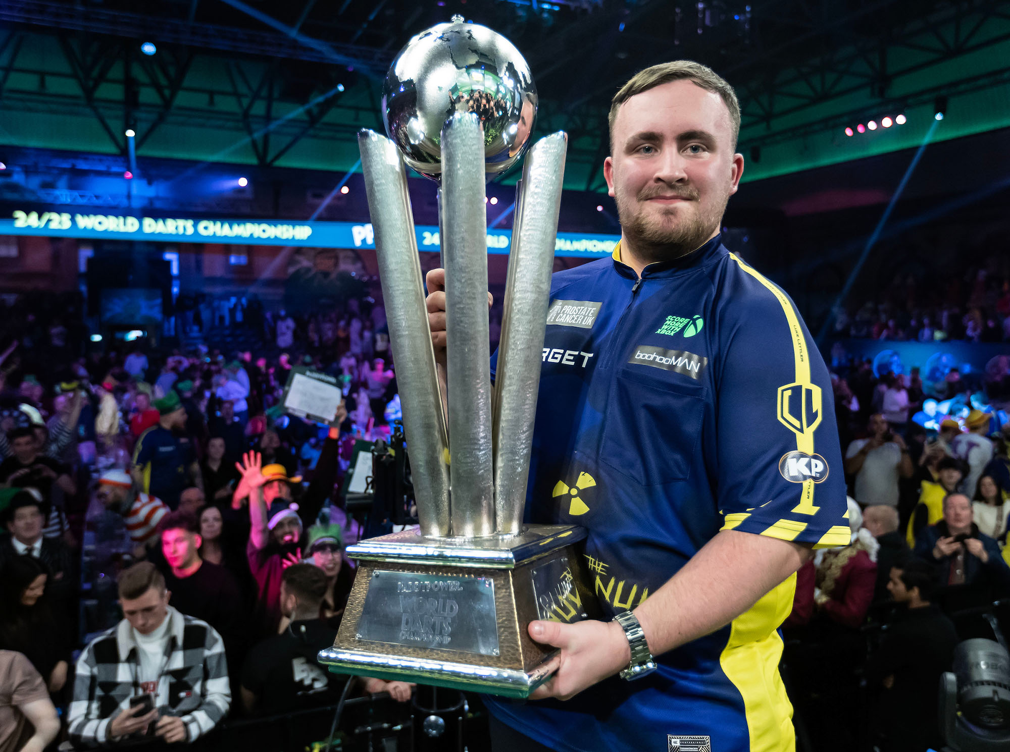 Luke Littler lifting the Darts World Championship trophy above his head, celebrating with a mix of disbelief and joy after becoming the youngest world champion in history.