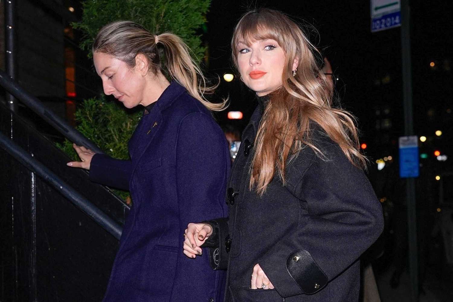 Taylor Swift and Este Haim walking through New York City at night, Taylor wearing a black Louis Vuitton coat, matching leather boots, and sheer tights, while Este wears a navy coat and black dress.