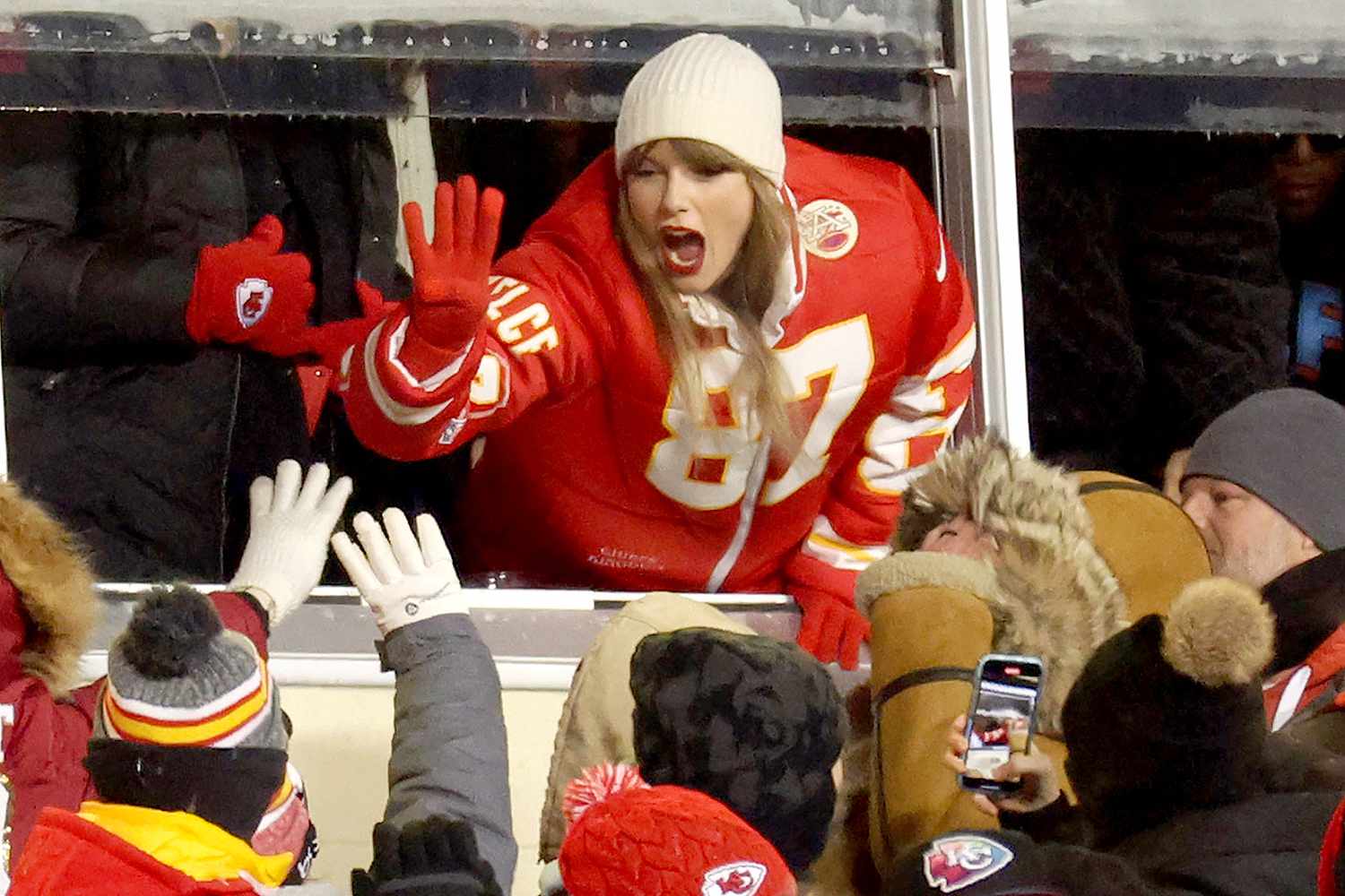 Taylor Swift high-fiving fans while wearing a Kansas City Chiefs jersey, smiling and engaging with the crowd.