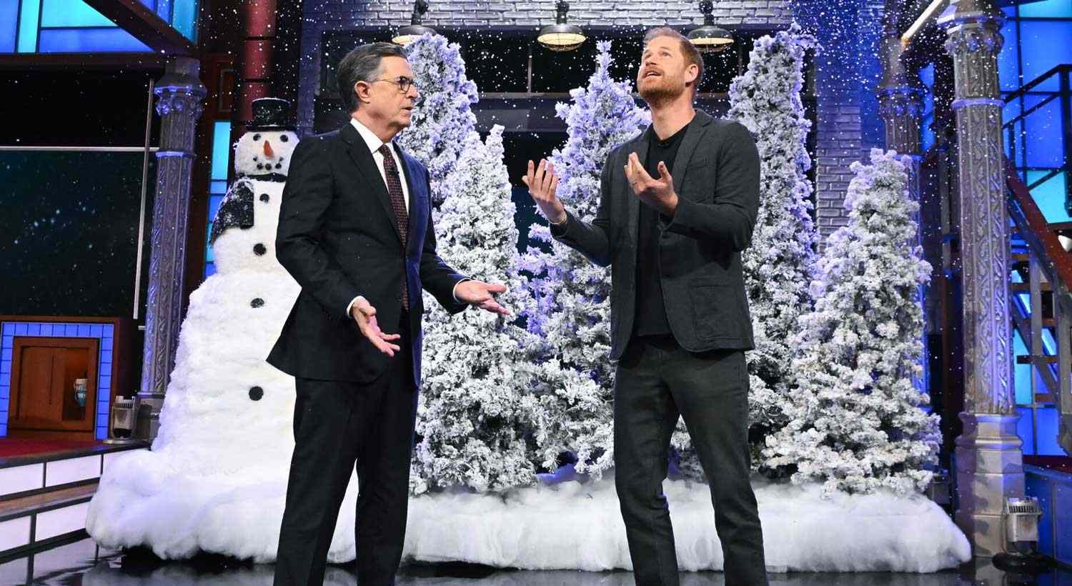 Prince Harry and Stephen Colbert acting out a festive Hallmark-style Christmas sketch on The Late Show, sharing an awkward laugh as artificial snow falls around them.