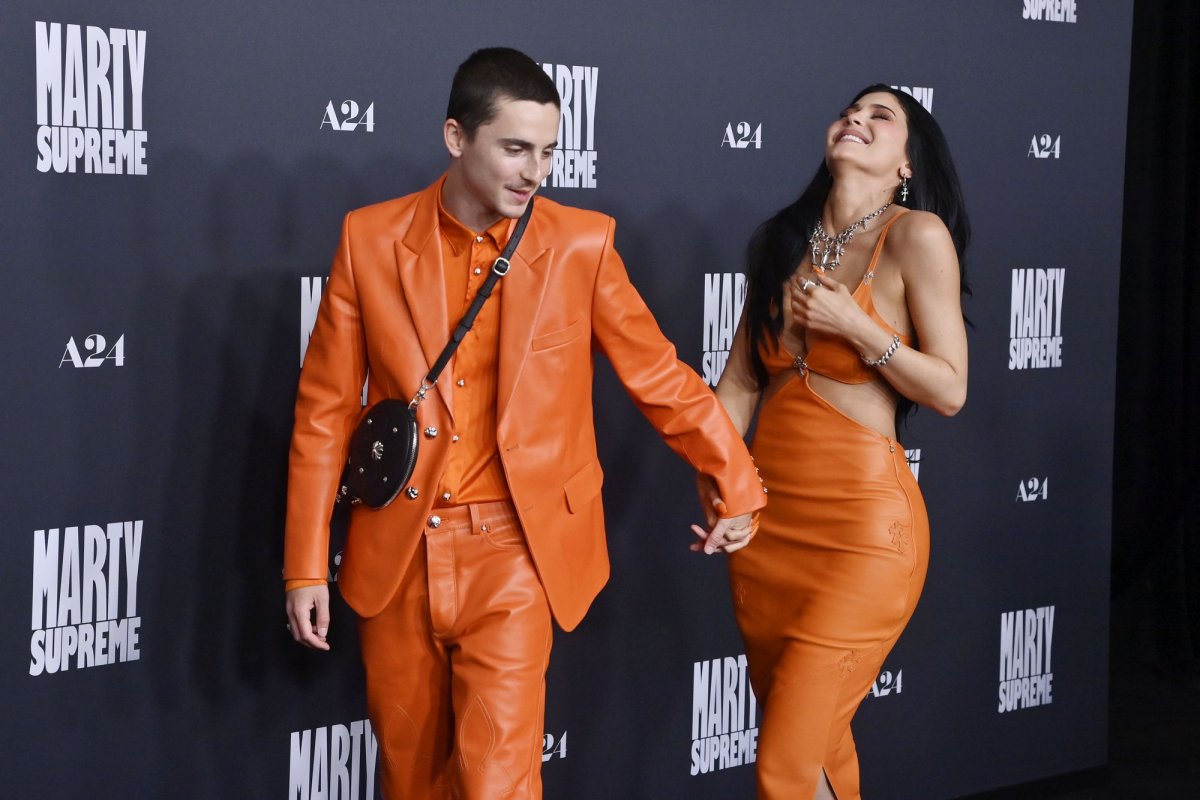 Kylie Jenner laughing as Timothée Chalamet tells a joke while they hold hands on the red carpet, both wearing coordinated bright orange outfits at the Marty Supreme premiere.