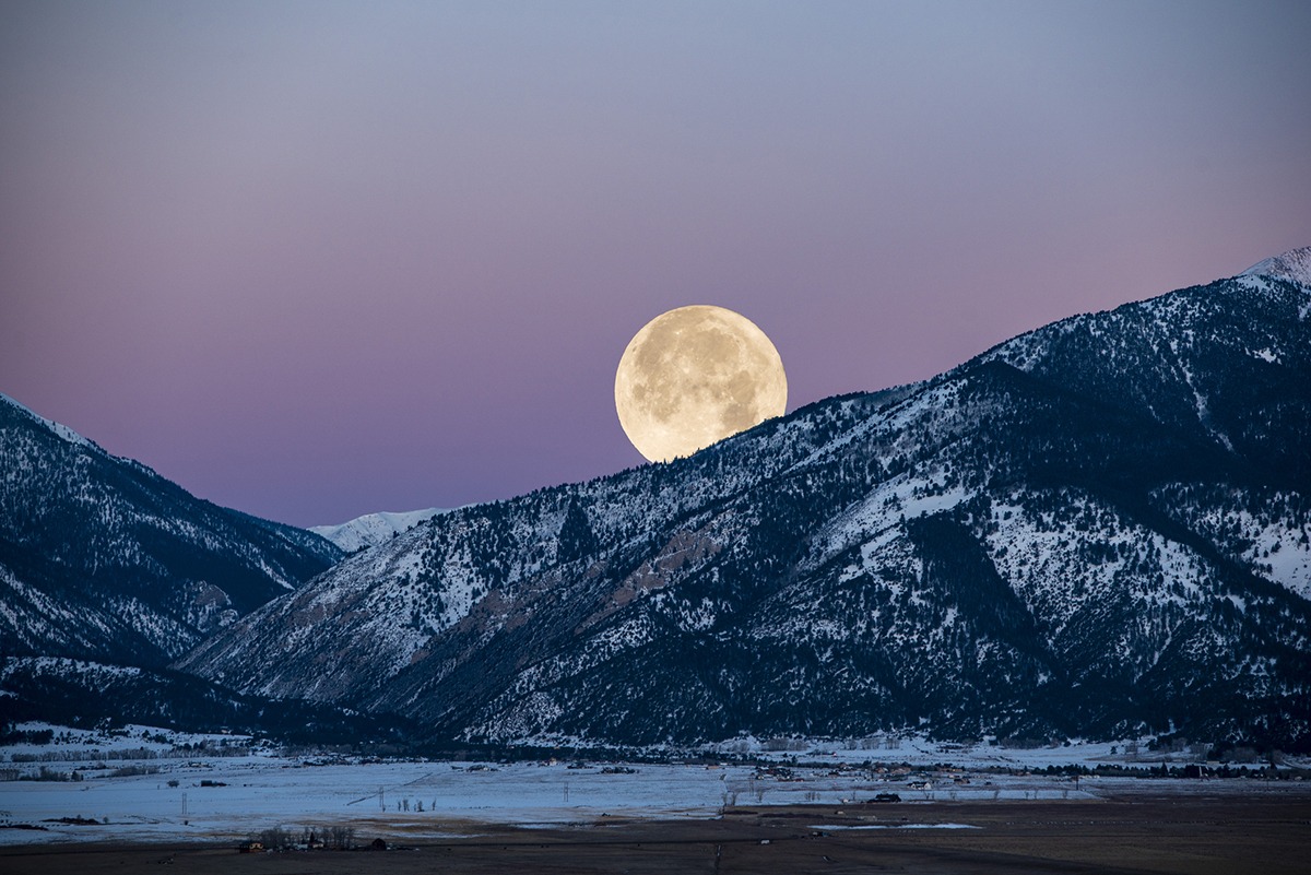 The supermoon rising over the crest of a mountain, its bright, oversized glow illuminating the landscape during the December Cold Moon.