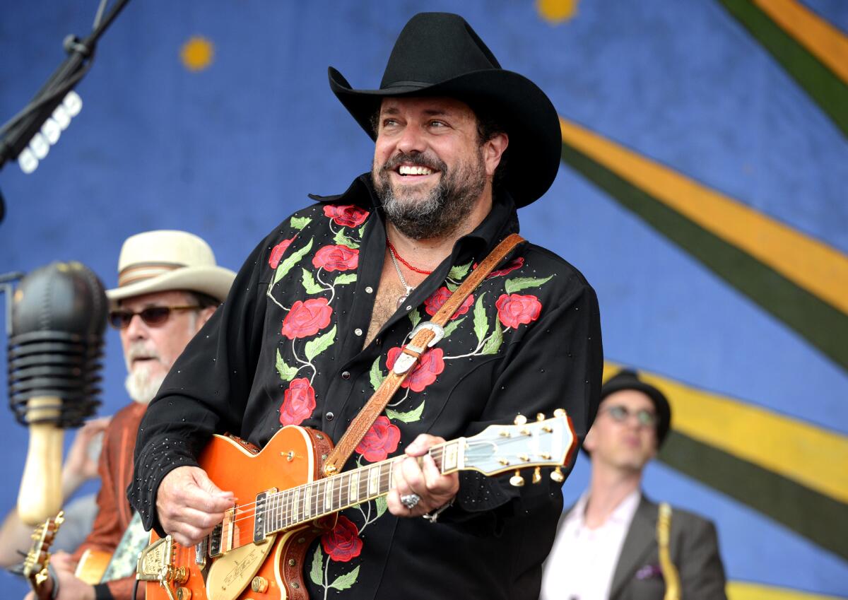 Raul Malo smiling while performing on stage, wearing a black shirt and holding a microphone, with colorful stage lights in the background.